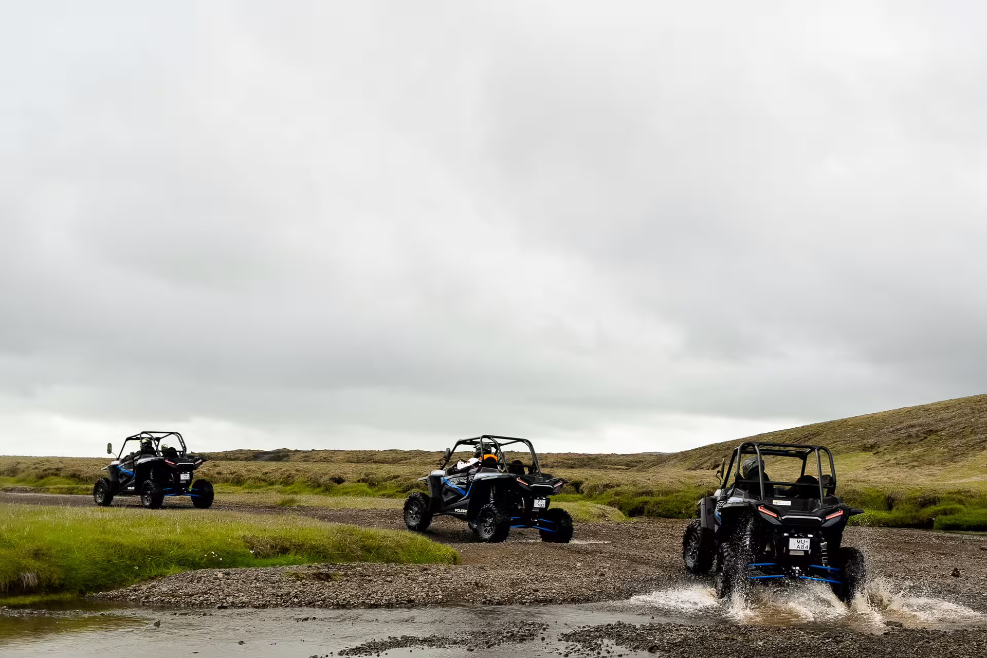 Three buggy UTVs driving through a shallow river crossing on Iceland's rugged highland terrain under overcast skies