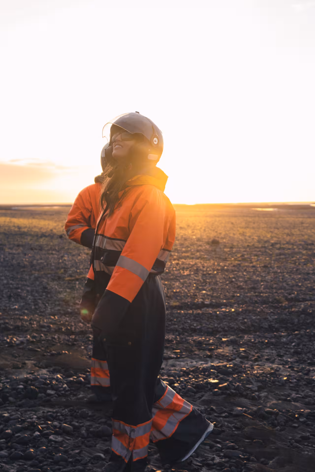 Person in orange hi-vis suit and helmet standing on black volcanic terrain at golden sunset in Iceland