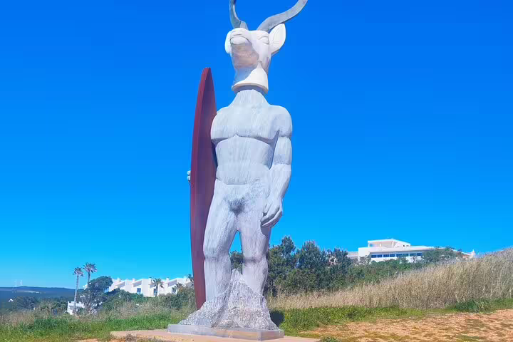 Sculpture of a bull-headed figure with surfboard under clear blue sky in Nazaré, Portugal.