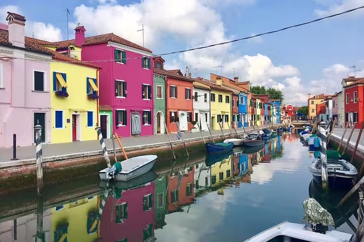 Colorful houses line the Burano canal, reflecting vibrant hues in the water on a serene Grand Canal boat tour.
