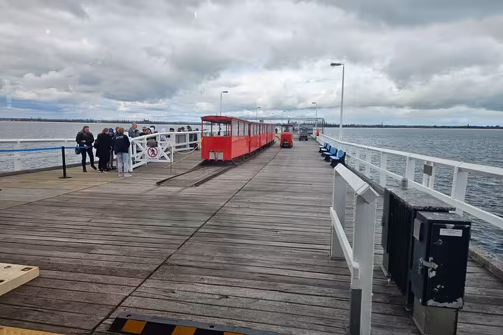 Tourists gather at Busselton Jetty while a red train awaits on the wooden pier under cloudy skies.