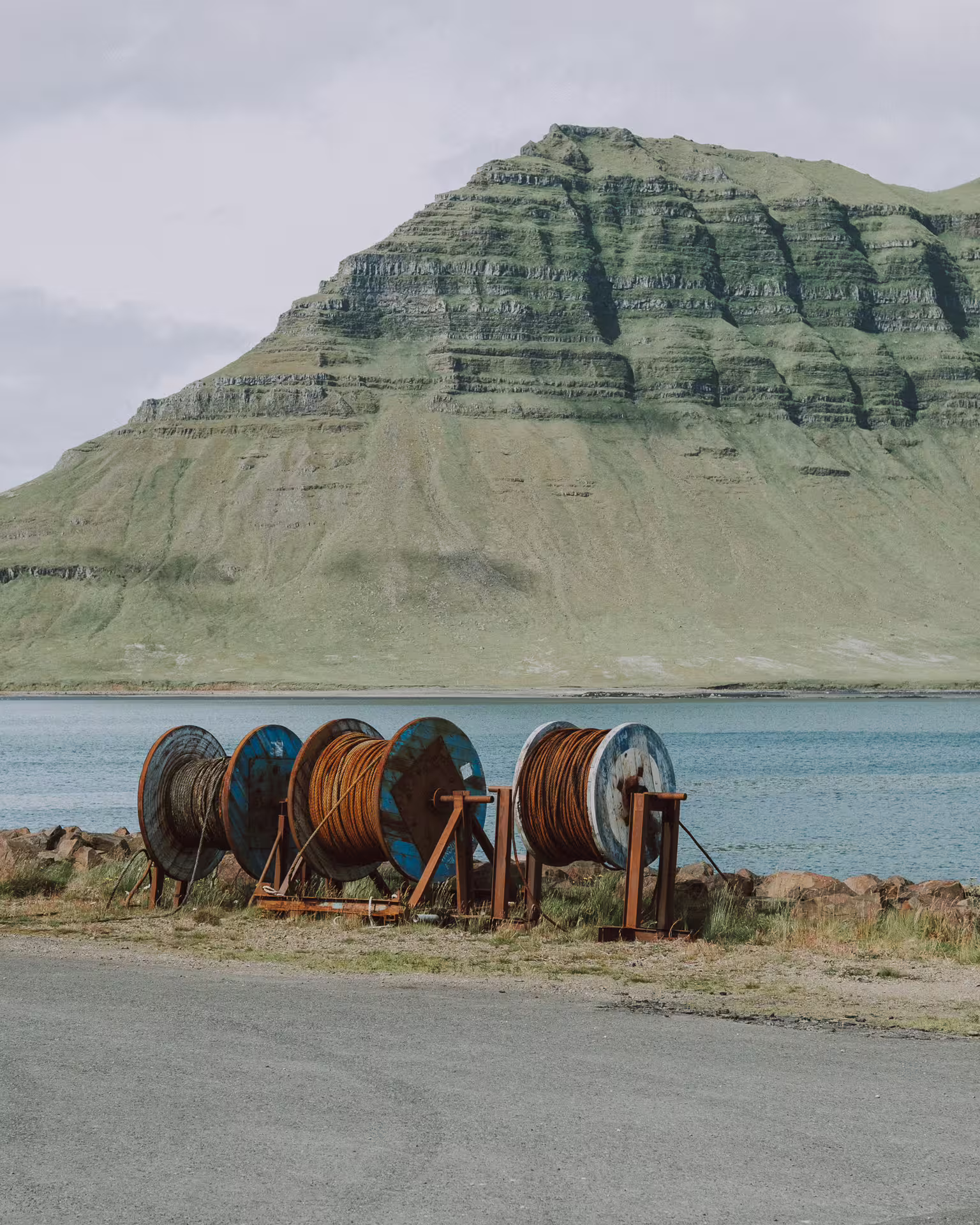 Large rusty cable reels by a fjord with a layered green mountain backdrop on the Snaefellsnes Peninsula