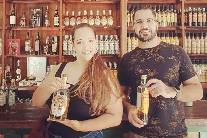 Two people smiling and holding bottles of cachaça in a traditional distillery during a tasting experience.