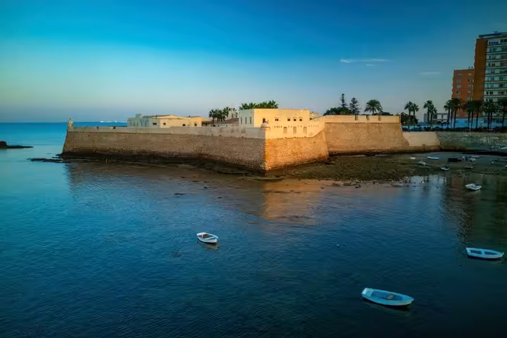 Historic Castillo de Santa Catalina fortress walls along Cadiz coastline at sunset with small boats in calm blue water