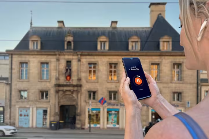 Woman holding smartphone with audio guide app in front of Hôtel d'Escoville in Caen, France