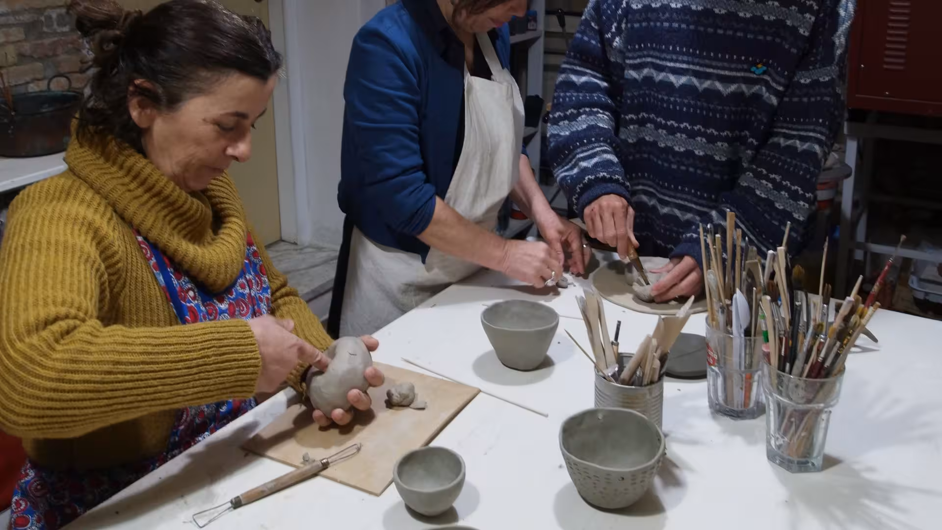 Three individuals sculpting clay in a cozy Cagliari ceramics workshop, with pottery tools and finished pieces adorning the table.