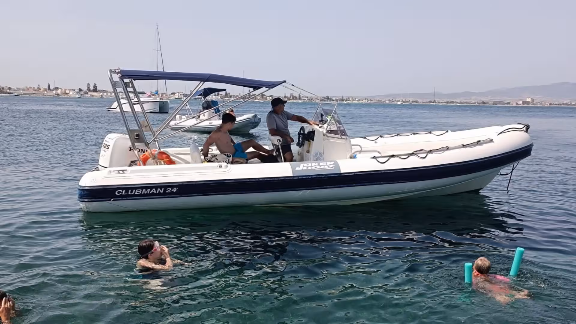 Tourists enjoying a Cagliari dinghy tour as they swim near a Joker Boat Clubman 24 under sunny skies.