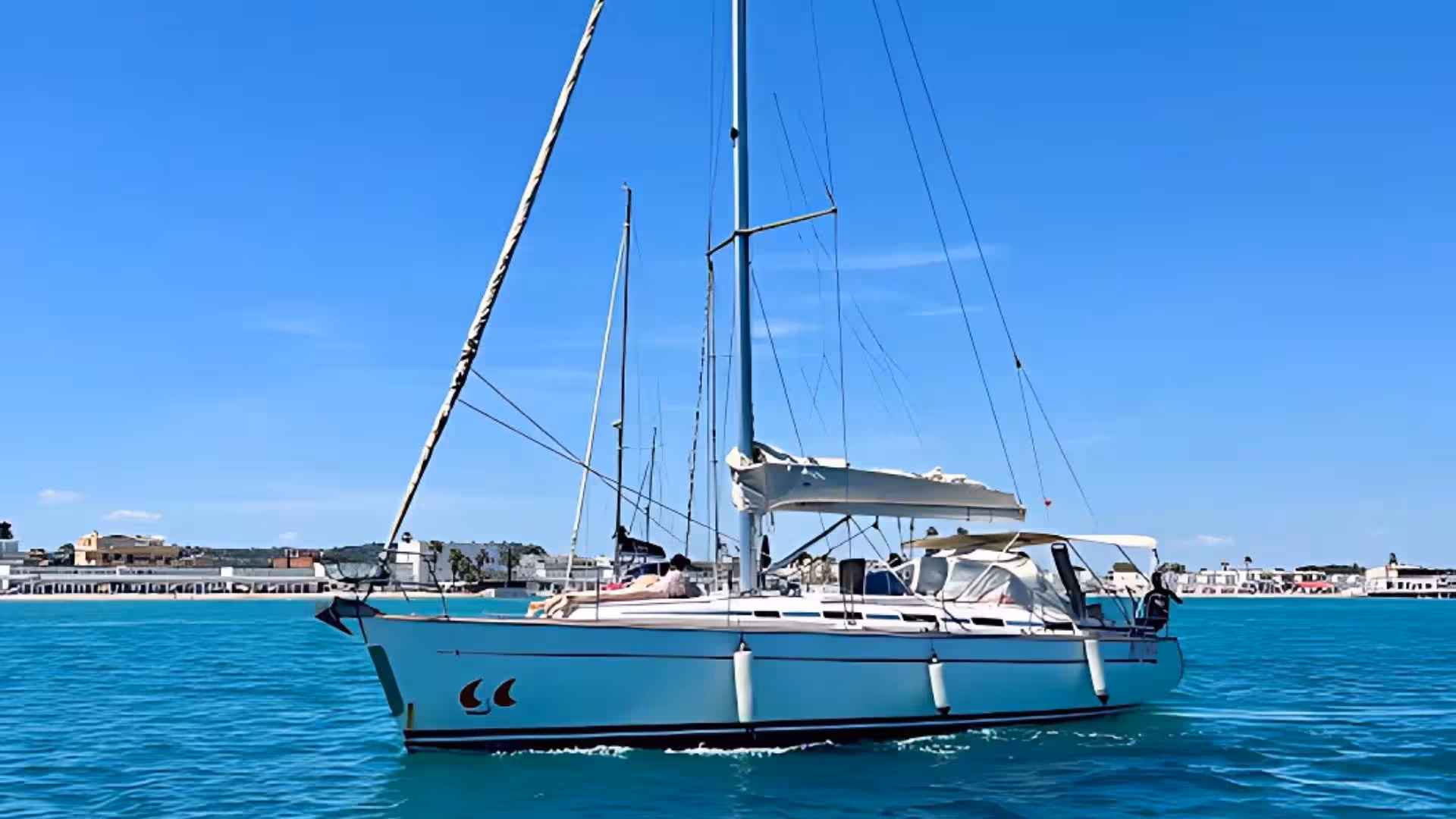 Elegant sailing boat cruising in the clear blue waters of Cagliari harbor, set against a backdrop of a vibrant marina.