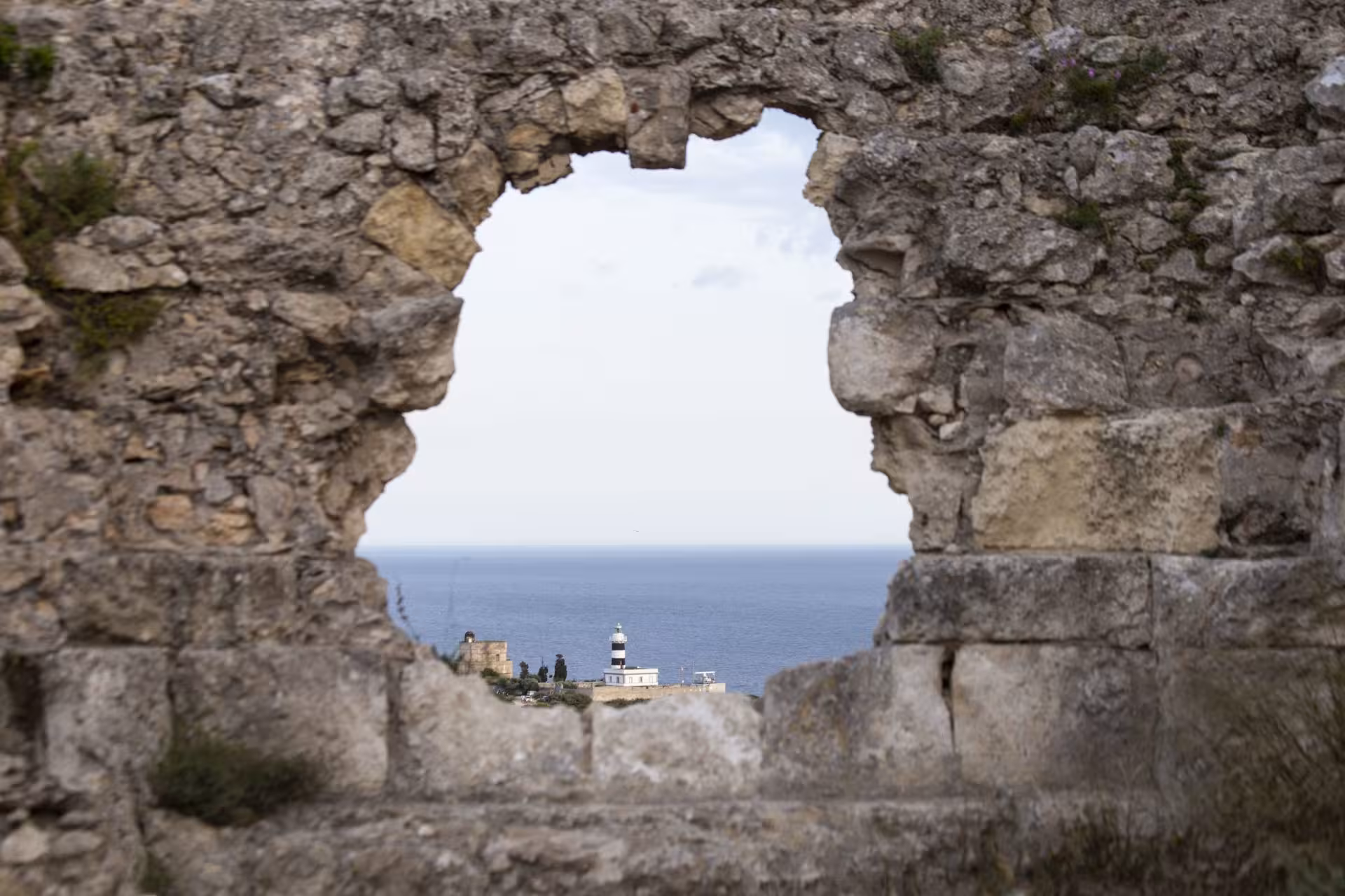 Ancient stone wall on Sant'Elia hill framing a distant lighthouse and sea view in Cagliari, Sardinia.