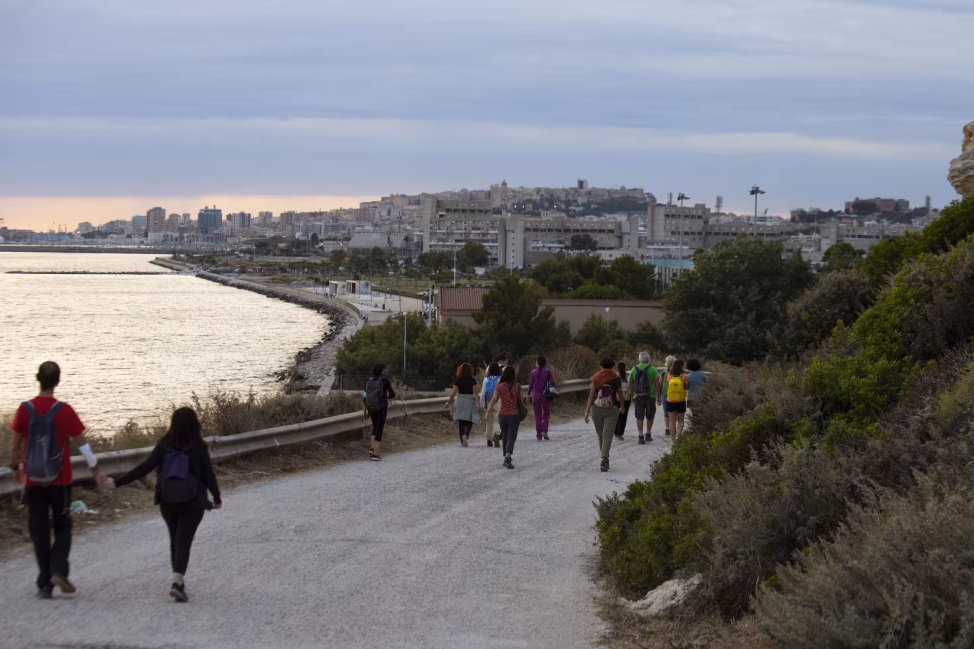 Hikers approaching Cagliari's coastline during sunset trek on Sant'Elia hill with city skyline in the background.