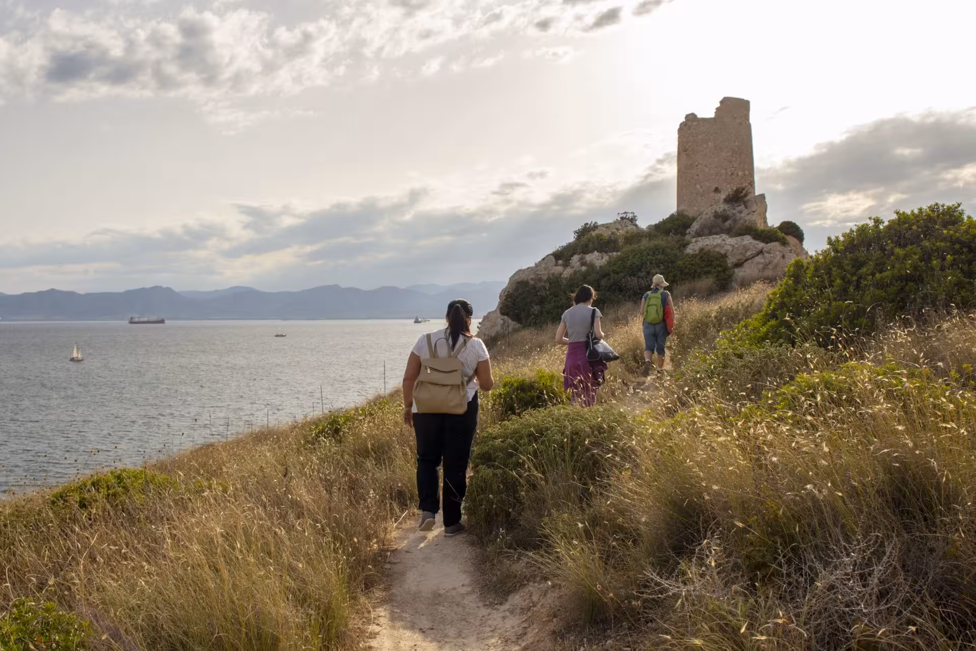 Group of hikers trekking towards a historic tower on Sant'Elia hill with panoramic sea views in Cagliari at sunset.