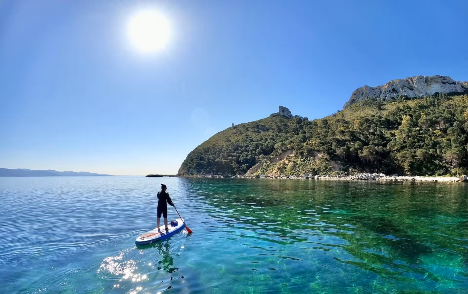 Solo paddleboarder on Cagliari's turquoise waters under a bright sun, with scenic view of Sella del Diavolo in distance.