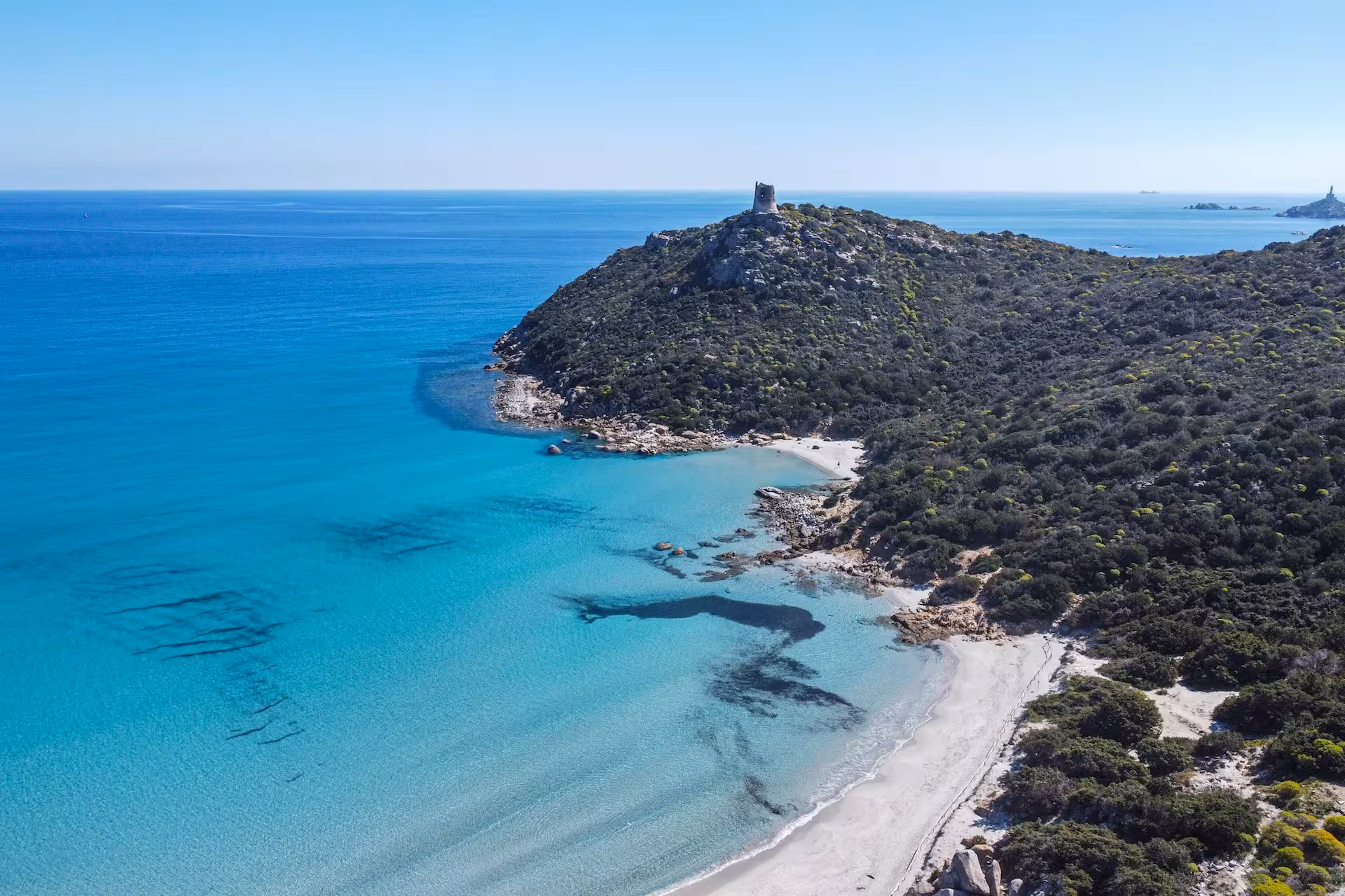 Aerial view of clear blue waters and a historic tower on a hill in Villasimius, Cagliari jeep tour destination.