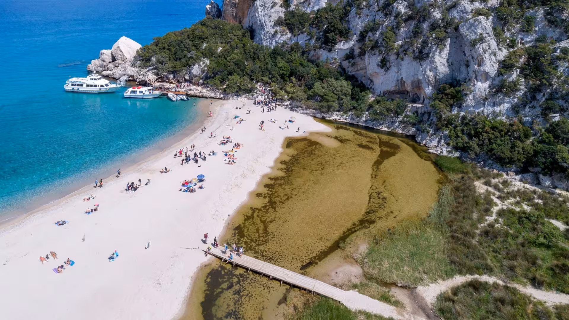 Aerial view of Cala Luna beach with white sand, lush cliffs, and boats anchored in crystal-clear blue waters.