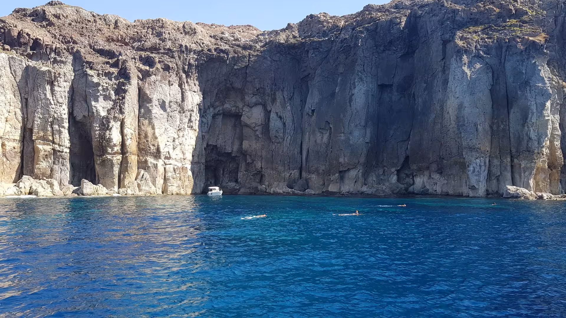 Swimmers exploring the crystal-clear waters and rugged cliffs near San Pietro Island on a boat tour.