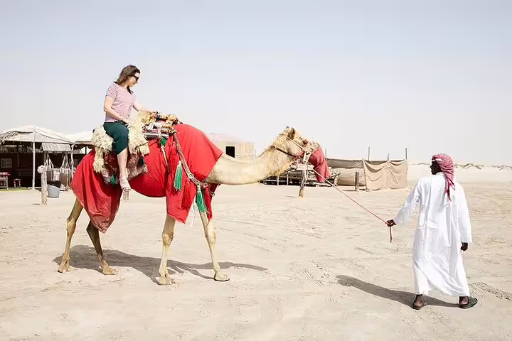 Tourist riding a camel led by a guide in traditional attire across the desert sands on a sunny day.