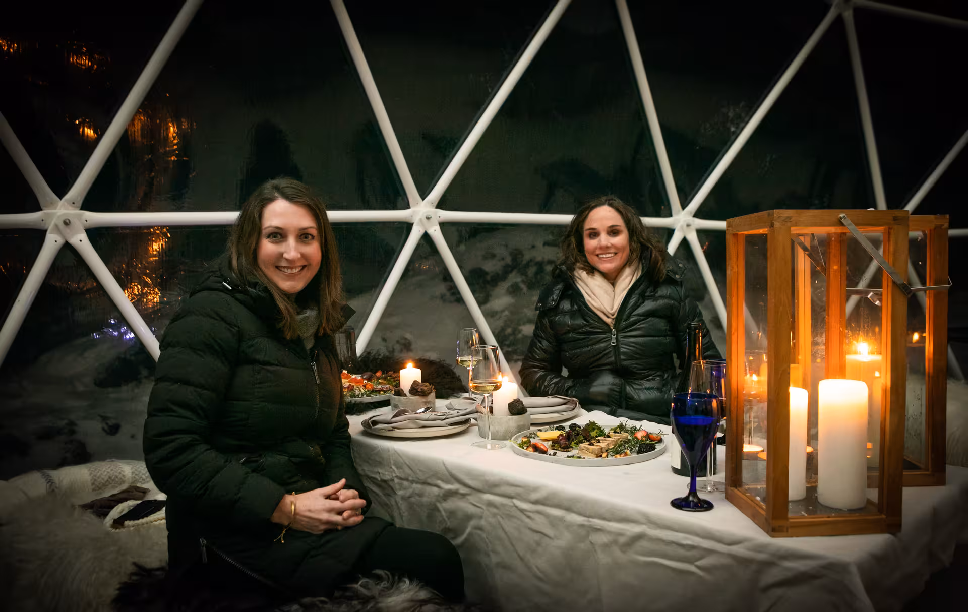 Two women enjoying a candlelit wine tasting and food pairing dinner inside a cozy glass geodesic dome by the lake