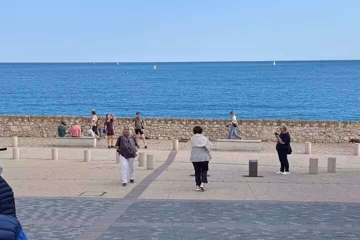 Tourists strolling along a sunny Mediterranean waterfront promenade with calm blue sea and sailboats in background