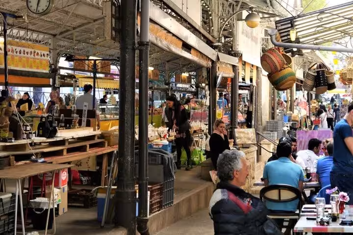 Bustling outdoor market with shoppers browsing vendor stalls under iron archways in a French town