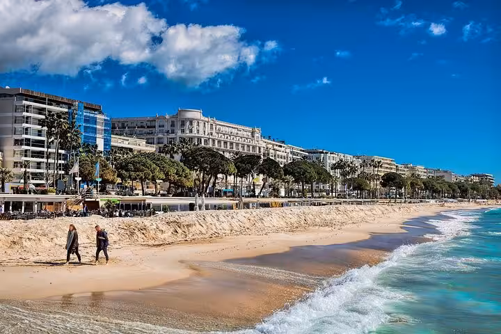 Sandy beach along Cannes promenade with palm trees, luxury hotels, and two people walking by turquoise sea