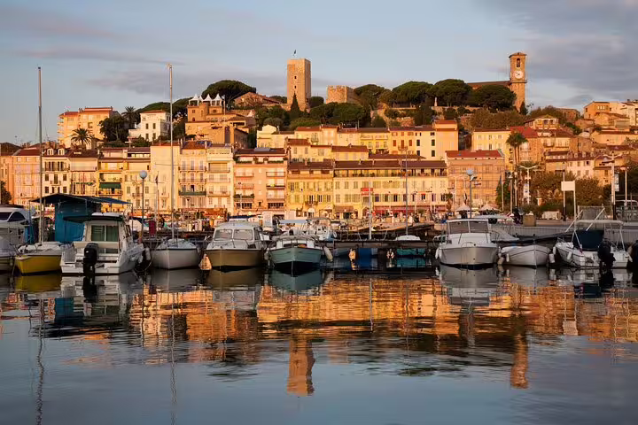 Cannes harbor at golden hour with boats moored and hilltop old town buildings reflected in calm water