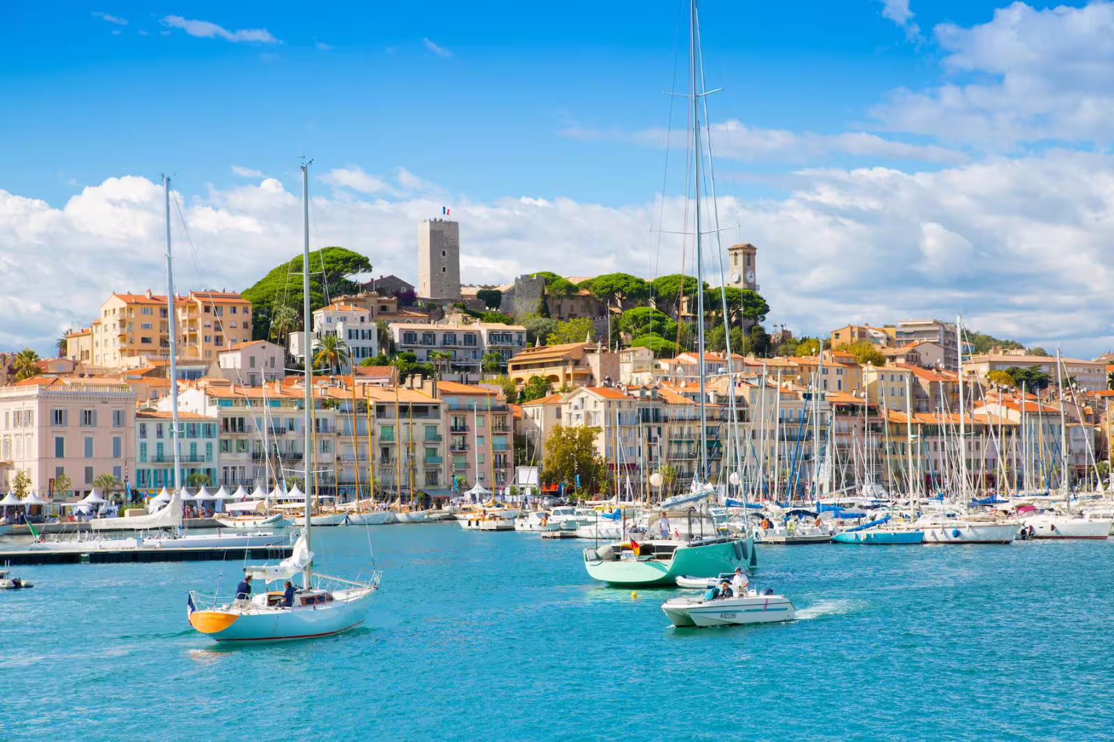 Cannes harbor with sailboats on turquoise water and colorful buildings on hillside, French Riviera
