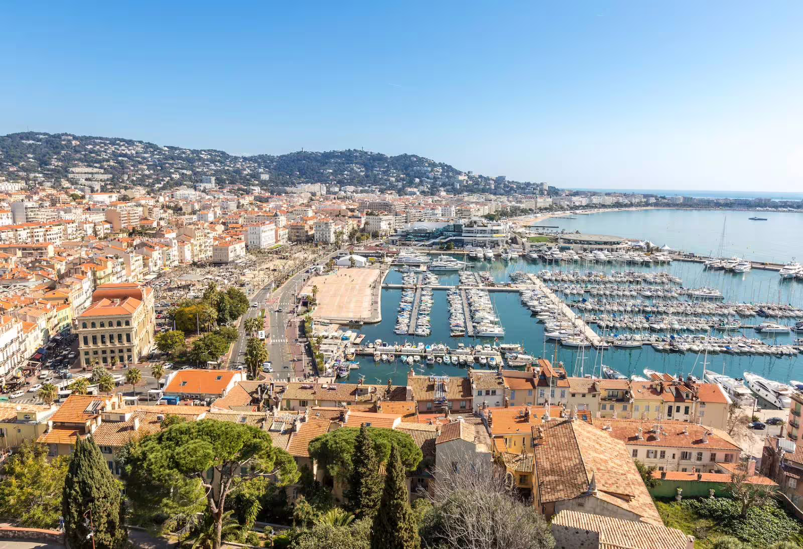 Aerial view of Cannes marina with yachts, terracotta-roofed buildings, and Mediterranean coastline on a sunny day