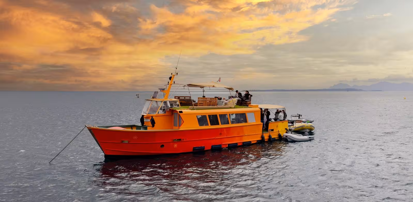 Vibrant orange party boat anchored in Cannes at golden sunset with guests enjoying the open deck experience.
