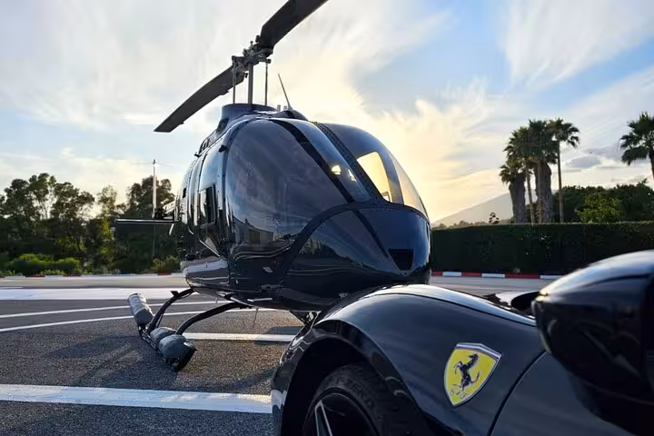 Black helicopter on helipad beside a Ferrari supercar with palm trees at sunset in Cannes