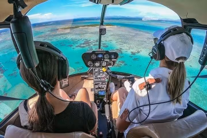 Two passengers in helicopter cockpit flying over turquoise coastal reef with headsets on
