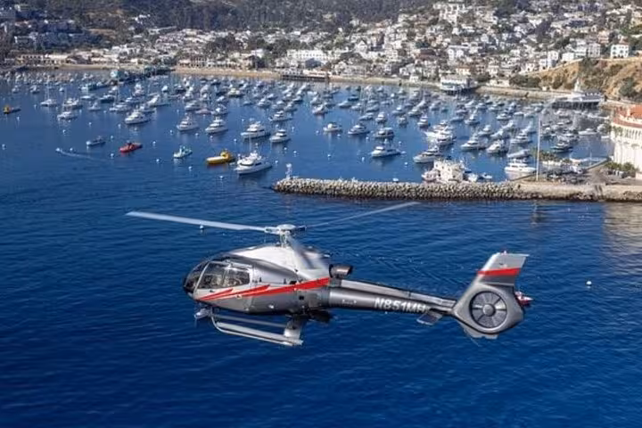 Silver helicopter flying over a Mediterranean harbor with boats and hillside town in background