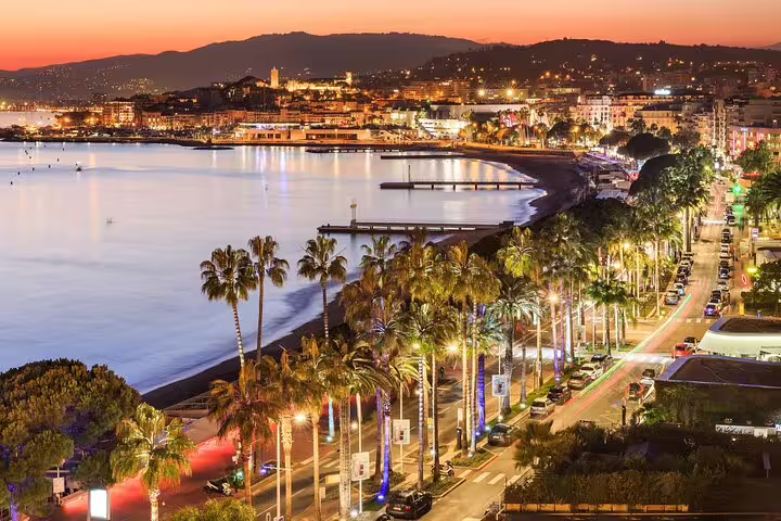 Cannes waterfront boulevard lined with palm trees at sunset, with glowing city lights reflecting on the Mediterranean Sea