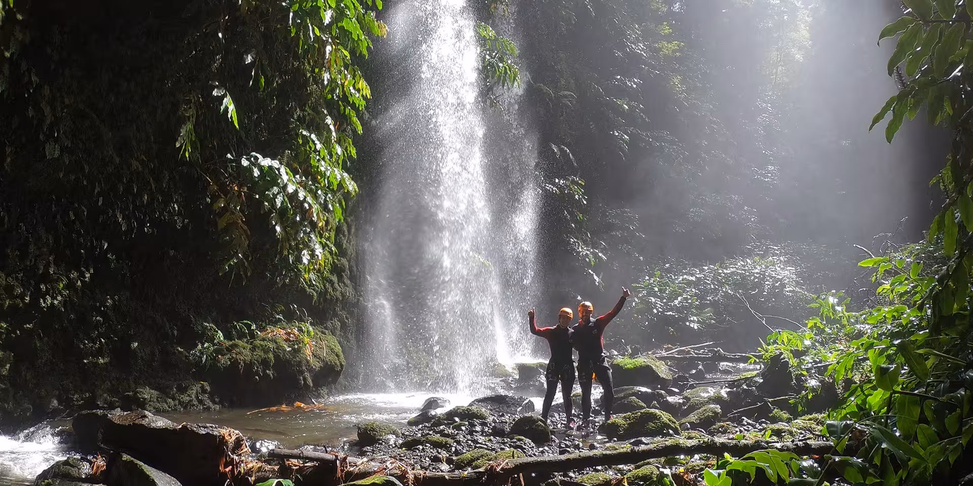 Canyoning adventurers in helmets celebrate by a rainforest waterfall on a guided canyoning experience tour
