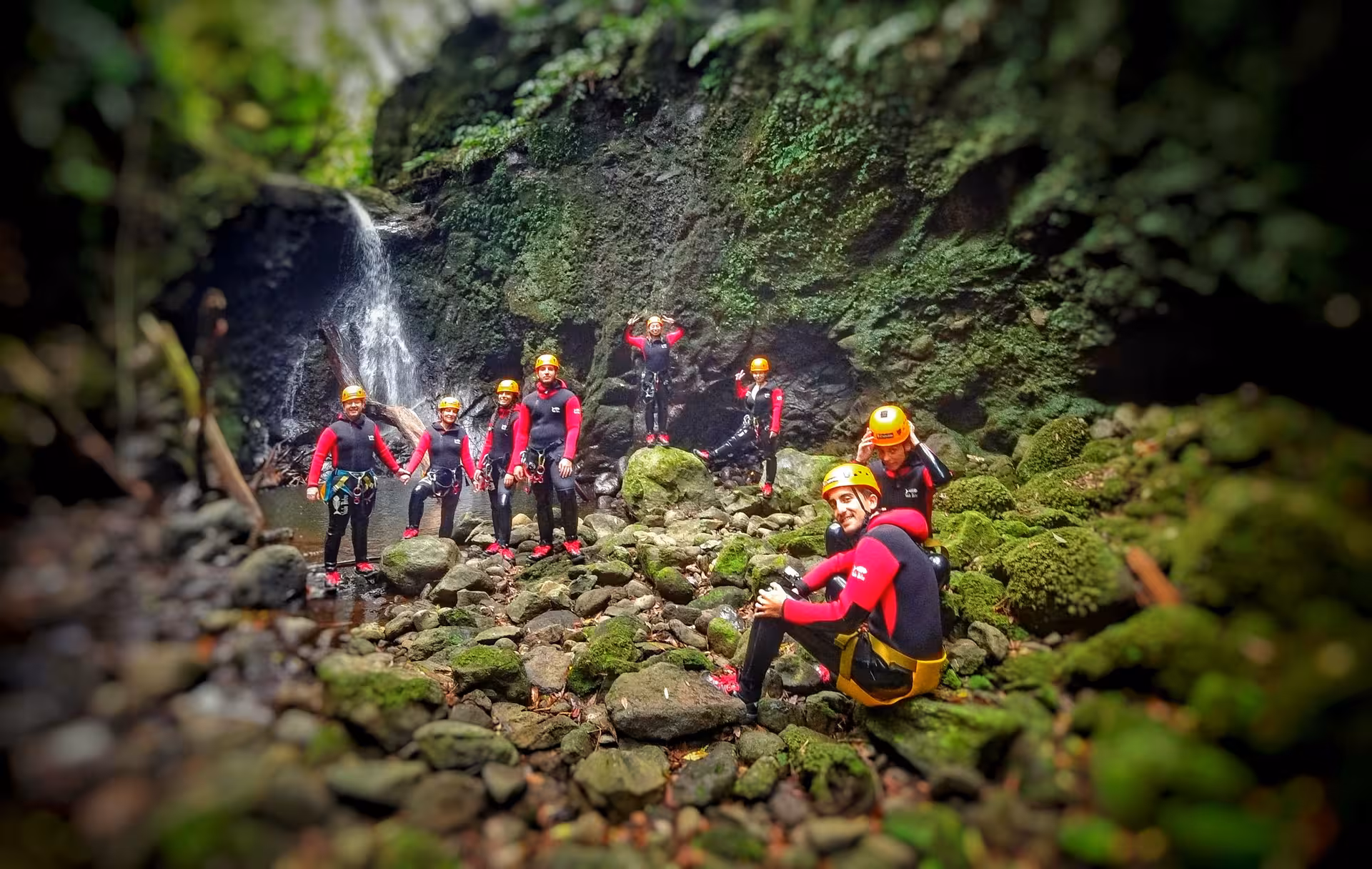 Canyoning group in Furnas Azores wearing wetsuits and helmets, exploring a mossy ravine with a small jungle waterfall