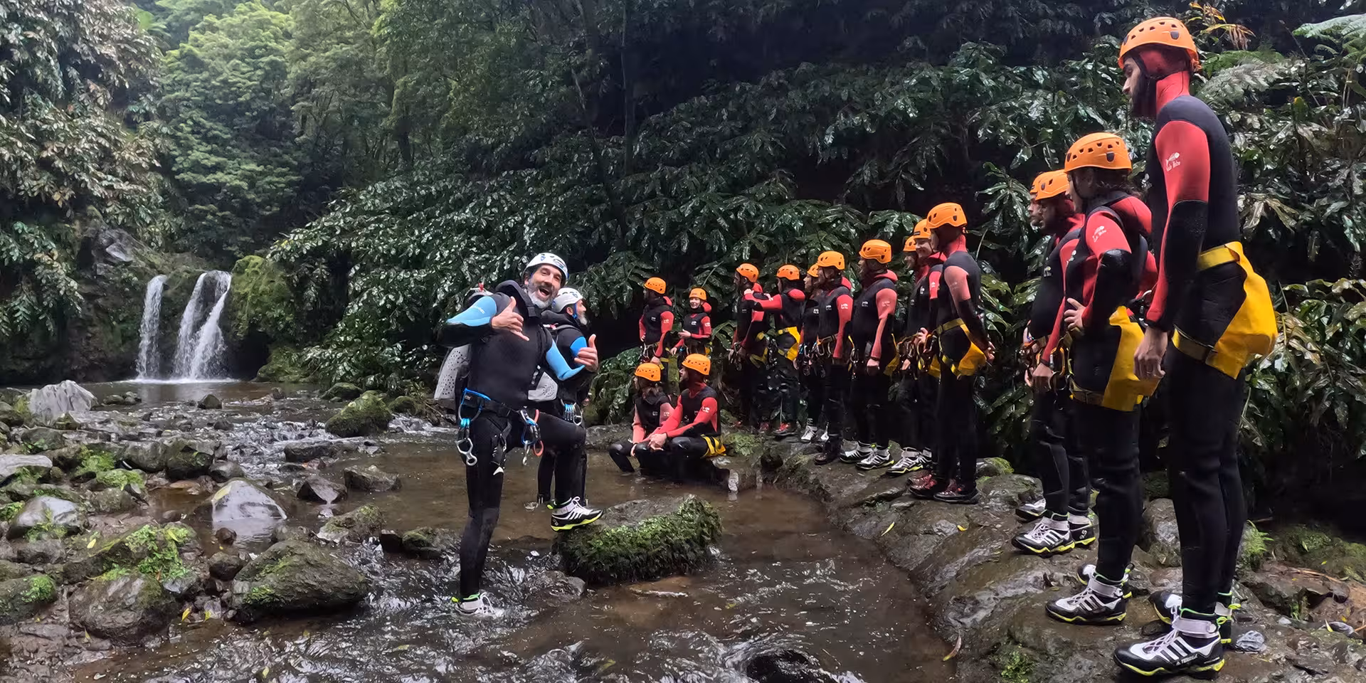 Canyoning group briefing by a waterfall in lush rainforest, wearing helmets and wetsuits for guided tour