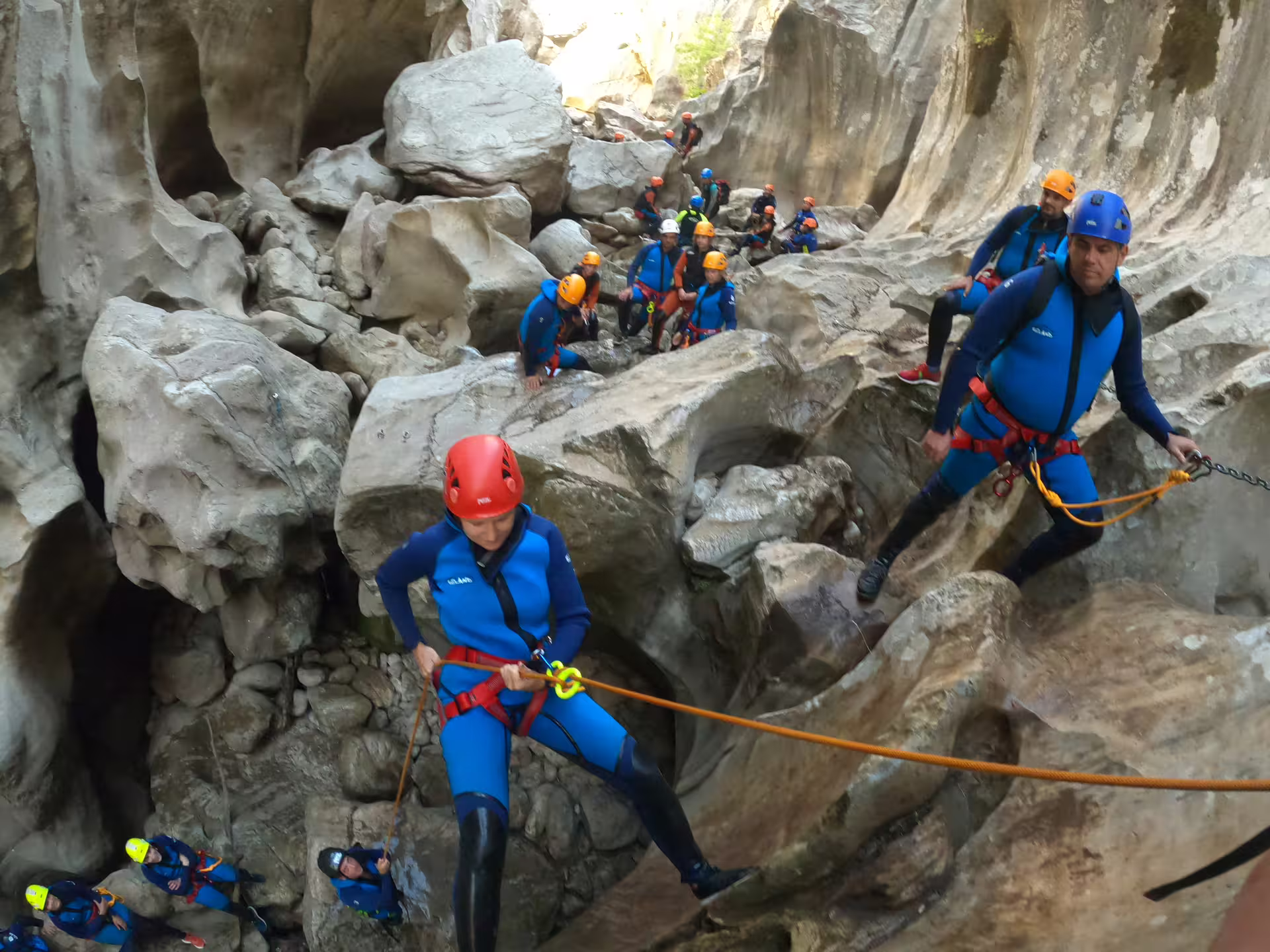 A group of canyoning tourists in blue wetsuits rappel down rugged rock formations at Las Buitreras Canyon, Málaga Province.