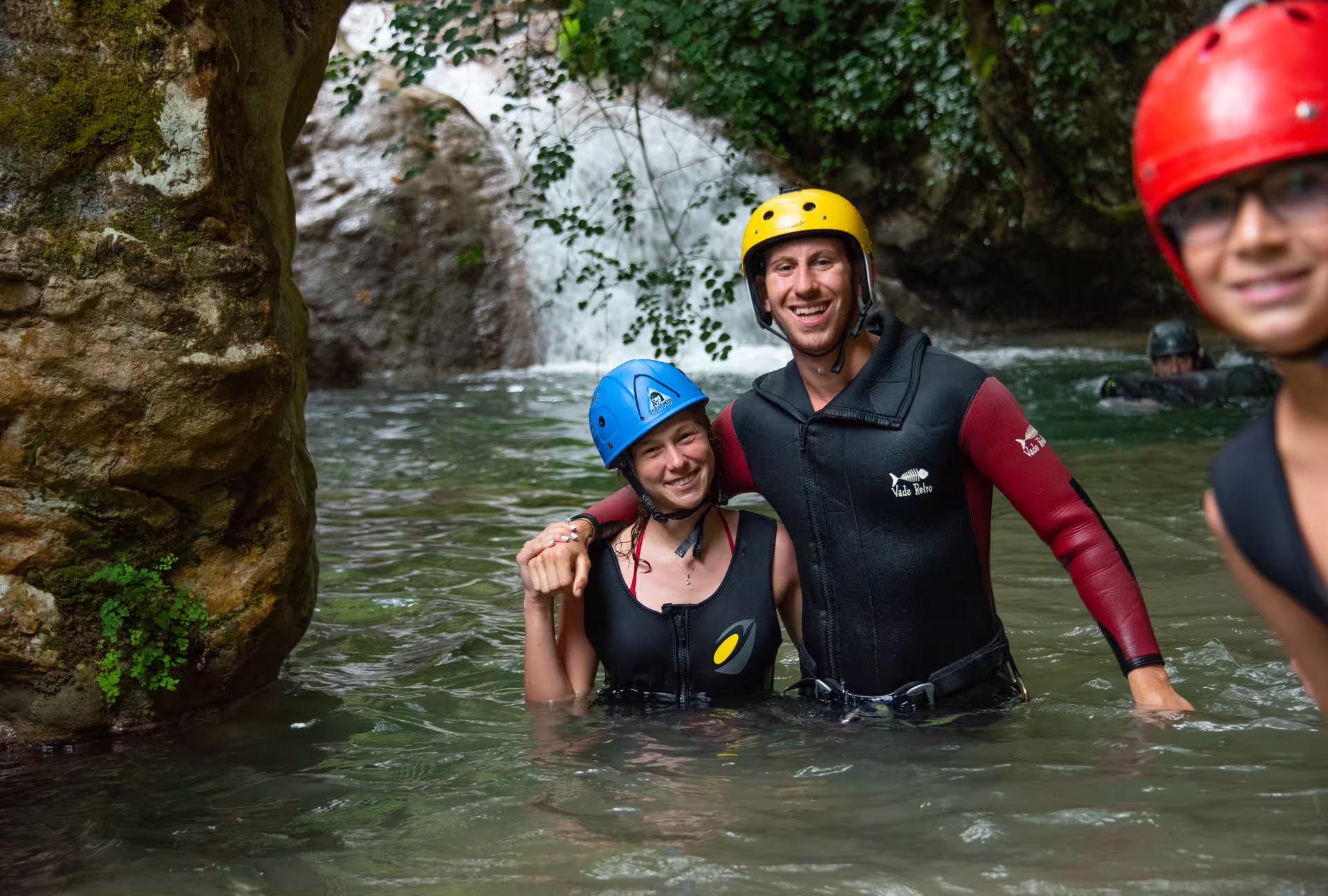 Smiling canyoning group in wetsuits and helmets wading in river pool at Neda Waterfalls canyon