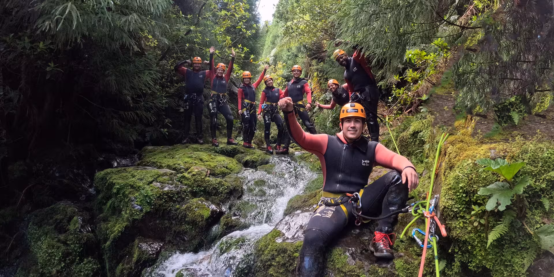 Canyoning Outward Shore group in wetsuits and helmets posing on mossy rocks by a rushing waterfall