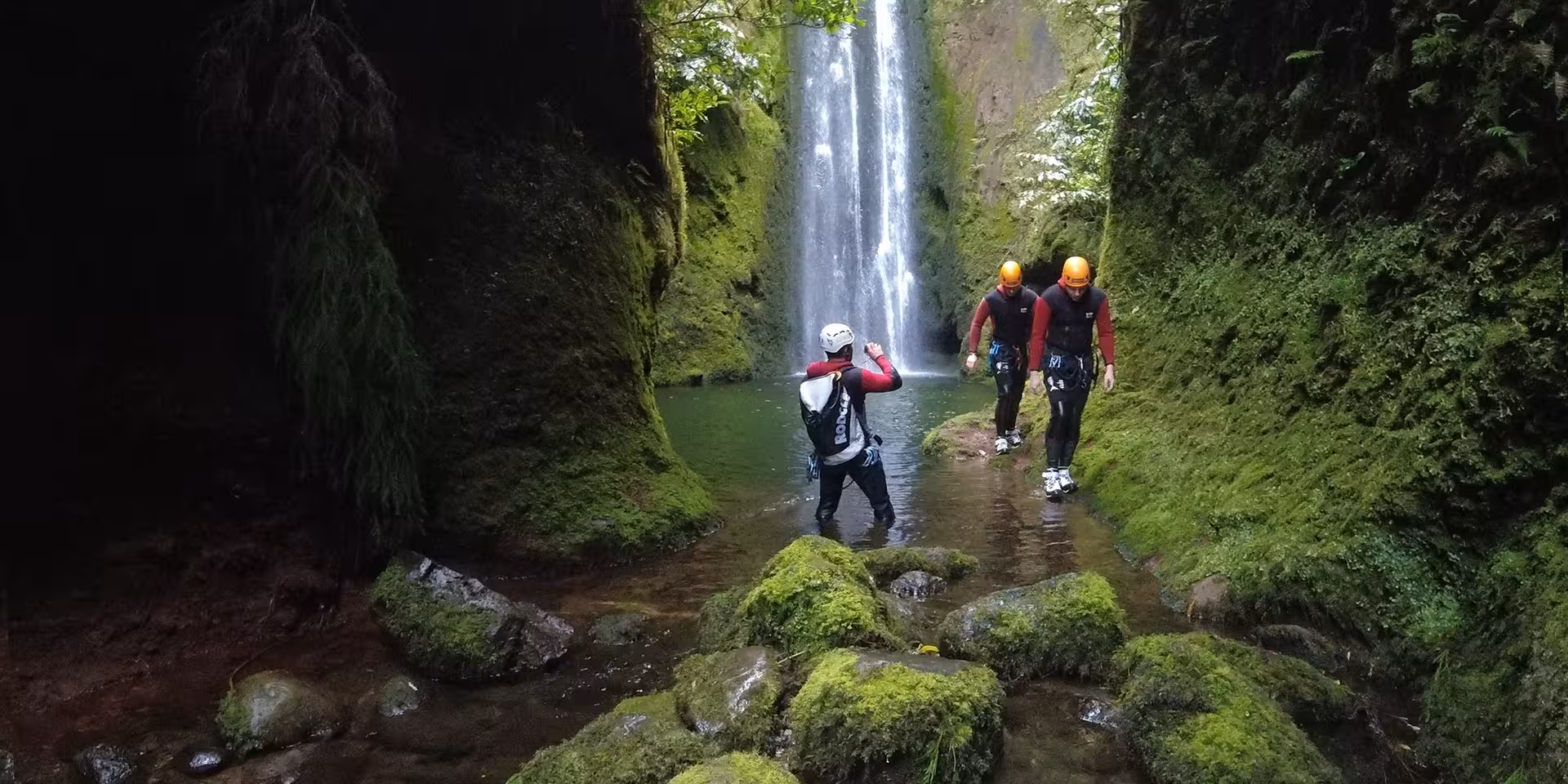 Canyoning Outward Shore group trekking through a mossy canyon stream toward a tall waterfall on a guided tour