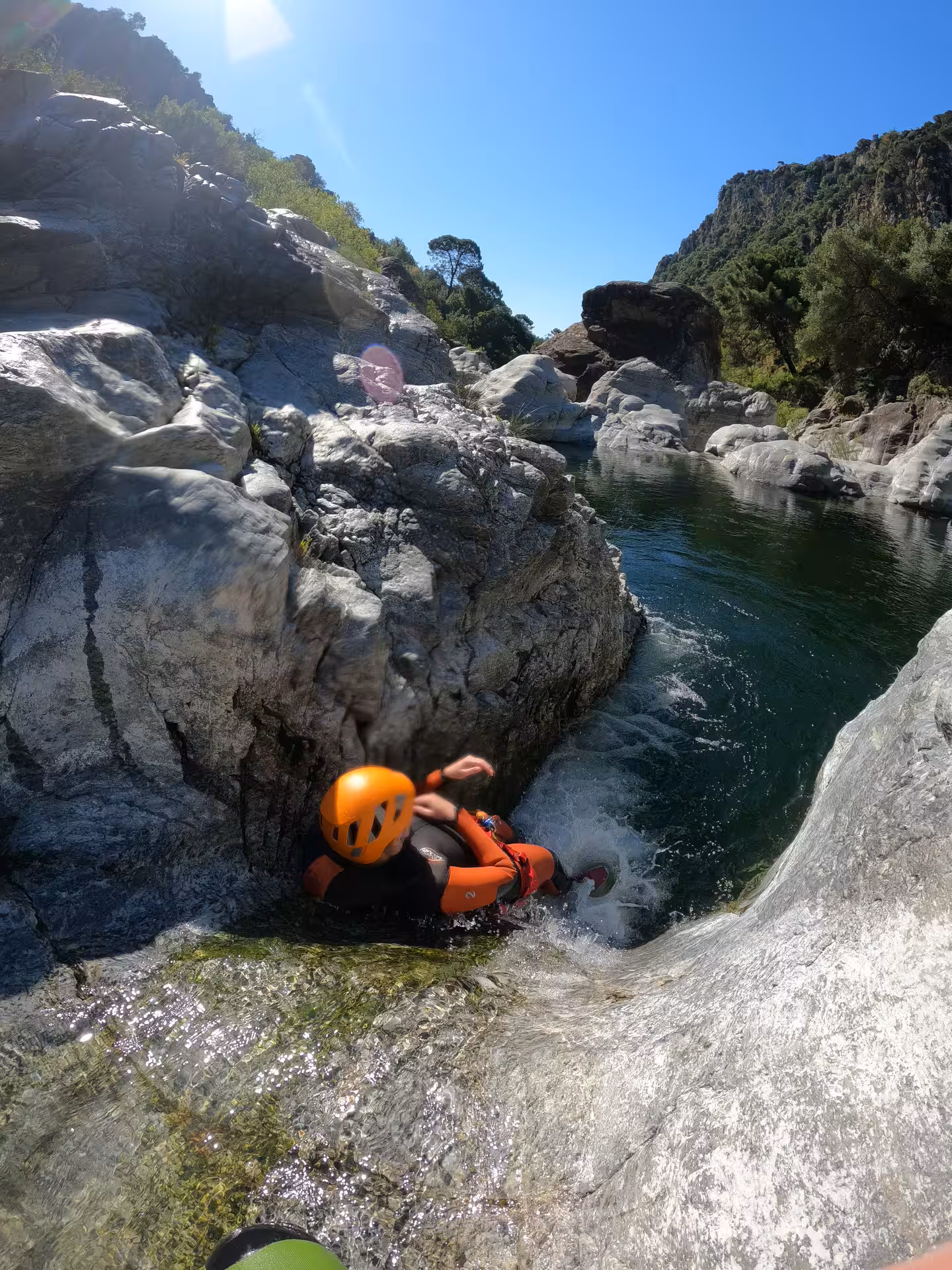 A helmeted adventurer slides down a natural rock chute into a crystal-clear pool during a guided canyoning tour in Guadalmina Canyon Estepona