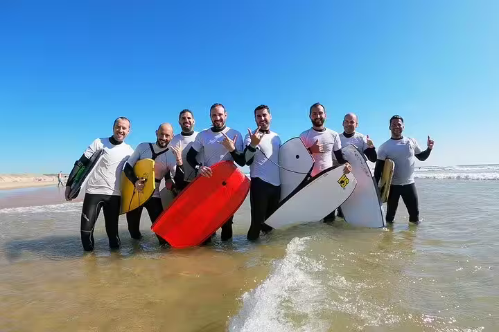 Group of happy bodyboarders in wetsuits enjoying Caparica beach adventure, holding colorful boards against a clear blue sky.