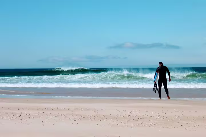 Person in wetsuit walking towards ocean waves with bodyboard, enjoying Caparica beach adventure under clear blue sky.