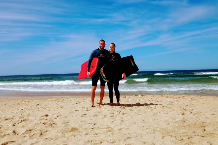 Couple with bodyboards on Caparica Beach ready for surf adventure under clear blue skies, perfect for yoga and surf enthusiasts.