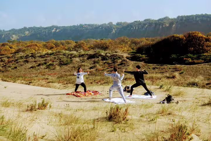 People practicing yoga on a sandy beach in Caparica with scenic coastal cliffs and greenery in the background.