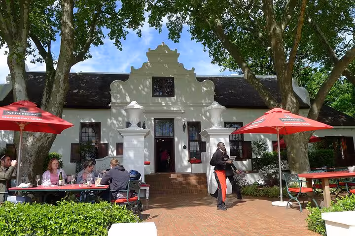 Historic Cape Dutch winery with red umbrellas, visitors dining outdoors, and lush surroundings in Cape Town's Winelands.