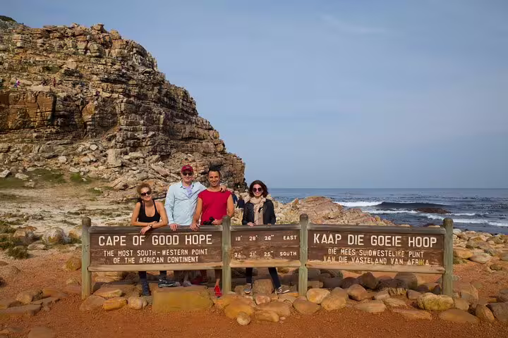 Four tourists pose by Cape of Good Hope sign with rocky cliffs and ocean backdrop on a guided tour from Cape Town.