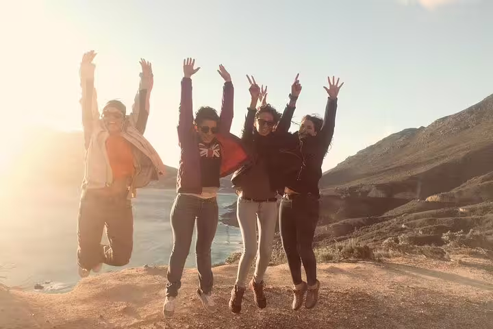Group of joyful tourists jumping with ocean and mountain backdrop at Cape of Good Hope, capturing adventure and excitement.