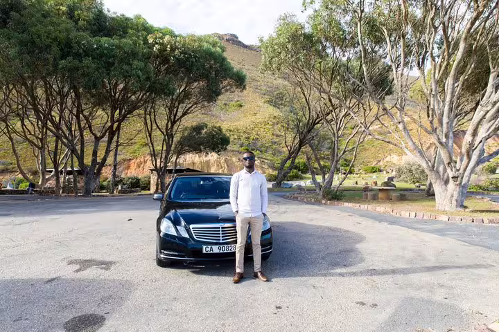 Tour guide standing in front of a black car at Cape of Good Hope parking area surrounded by trees.