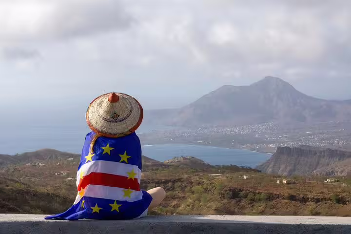Traveler wrapped in Cape Verde flag enjoys panoramic view of Santiago Island's mountains and coastline.
