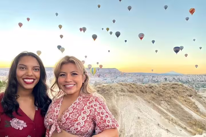 Two smiling women enjoy a Cappadocia hot air balloon experience, surrounded by a stunning landscape at dawn.